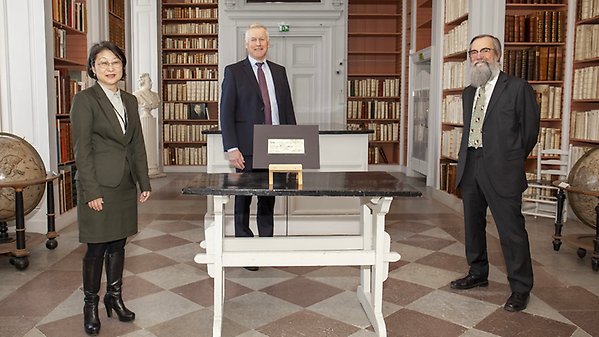 Professor Coco Norén, Professor Anders Malmberg and Library Director Professor Lars Burman standing around a table where a manuscript is displayed. Bookshelves in the background.